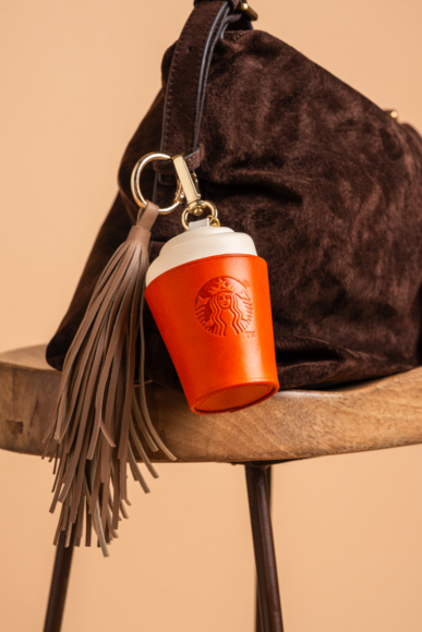 Starbucks The Autumn Charm - orange leather coffee-cup bag charm with a tan leather tassel clipped to a brown suede handbag, made by The Robin Collective